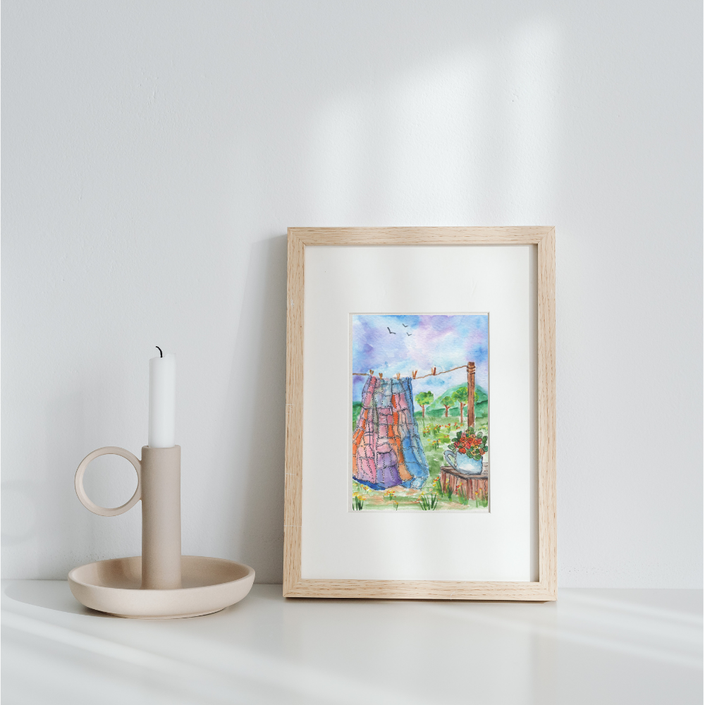 Framed watercolor quilt on a clothesline and teapot filled with flowers print leaning against wall with candleholder