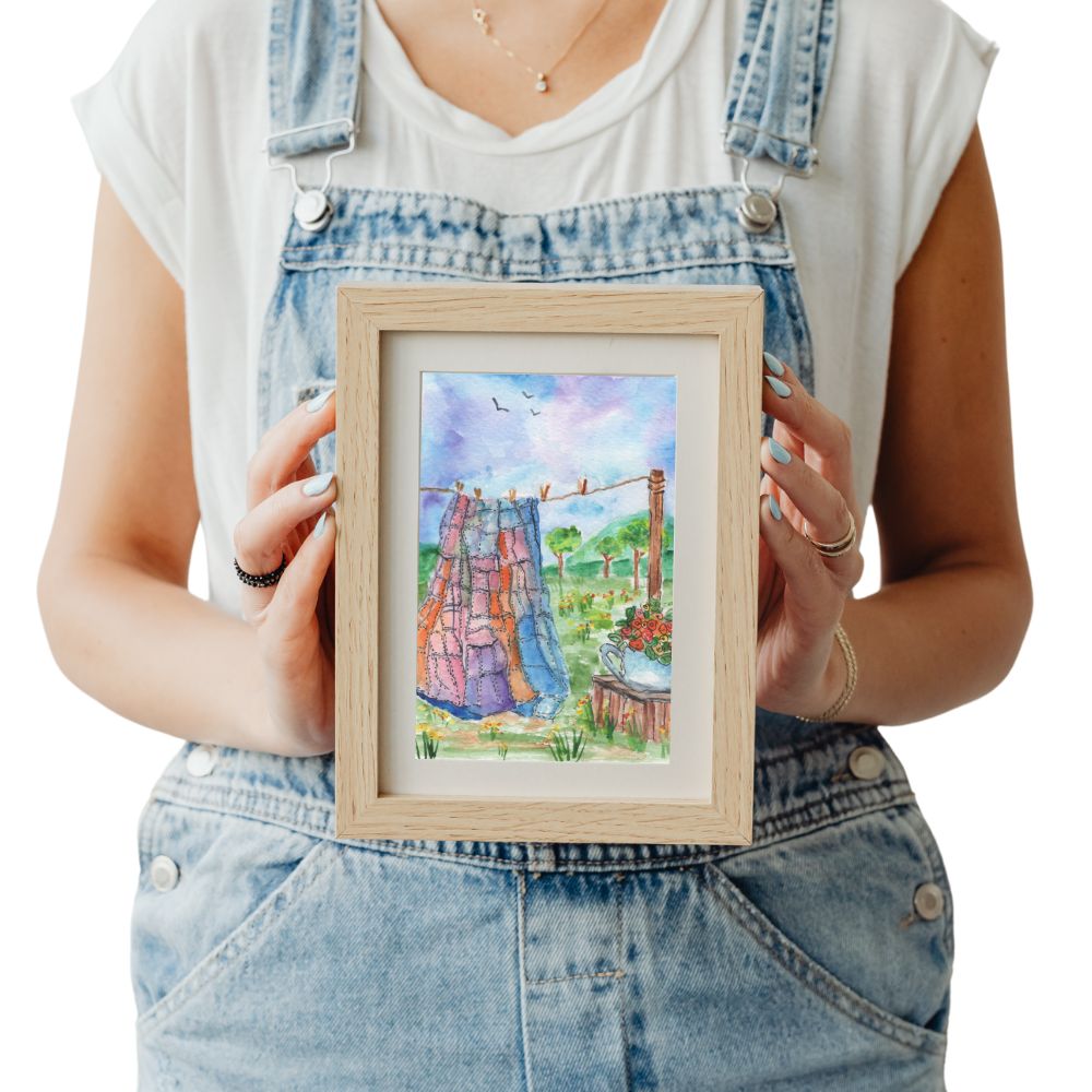 Woman holding framed quilt art print featuring a quilt on a clothesline, teapot and birds