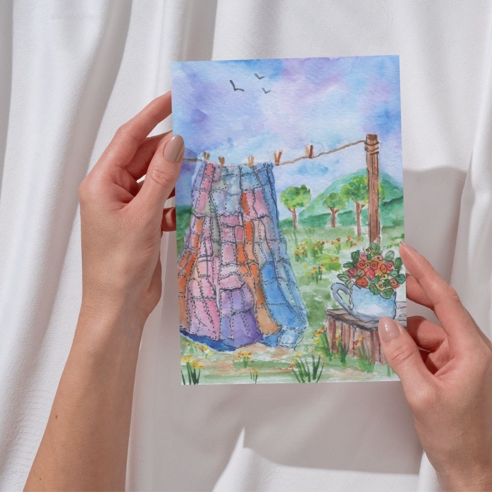 Woman holding a watercolor greeting card with quilt and floral teapot scene