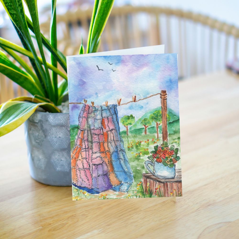 Lifestyle image of a quilt-themed greeting card leaning on a table with a plant